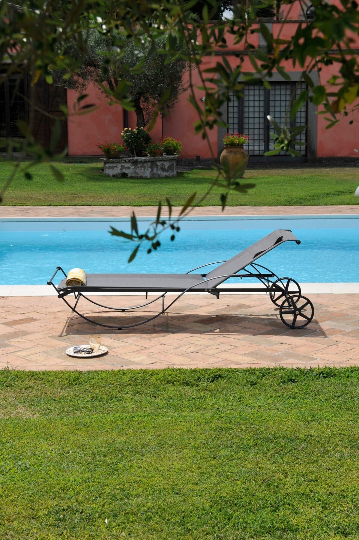 Eine Szene im Freien mit einem Liegestuhl in grafitgrau mit angenehmer Bespannung neben einem Pool. Auf dem Stuhl steht ein Sonnenhut und daneben auf dem Boden ein Teller mit Erfrischungen. Im Hintergrund ist ein Gebäude mit großen Fenstern und Topfpflanzen zu sehen. Bäume rahmen die ruhige, von der Aurora beleuchtete Szene ein.