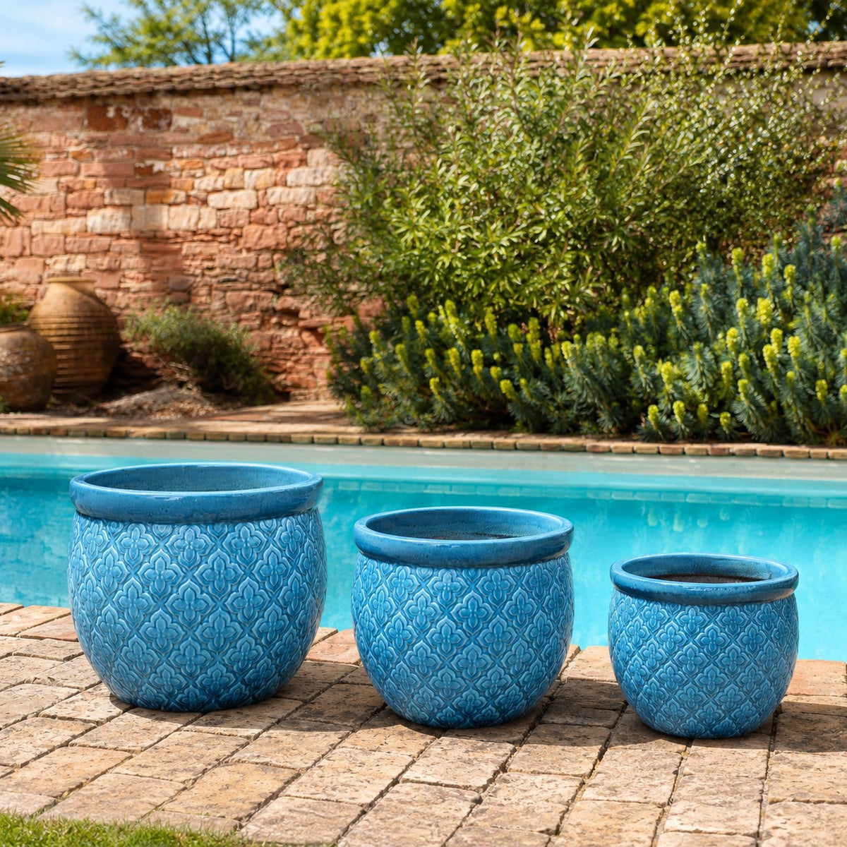 Drei blaue Keramikblumentöpfe unterschiedlicher Größe stehen nebeneinander auf einer Steinterrasse am Swimmingpool, mit einer Ziegelmauer und Gartengrün im Hintergrund.