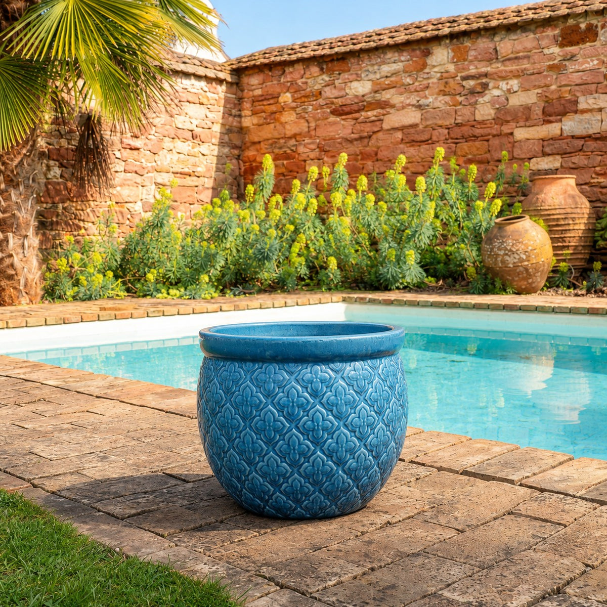 Ein großer blauer dekorativer Übertopf steht an einem sonnigen Tag auf einer Steinterrasse neben einem klaren Swimmingpool, mit Grünpflanzen, einer Palme, einem rustikalen Terrakottatopf und einer Steinmauer im Hintergrund.