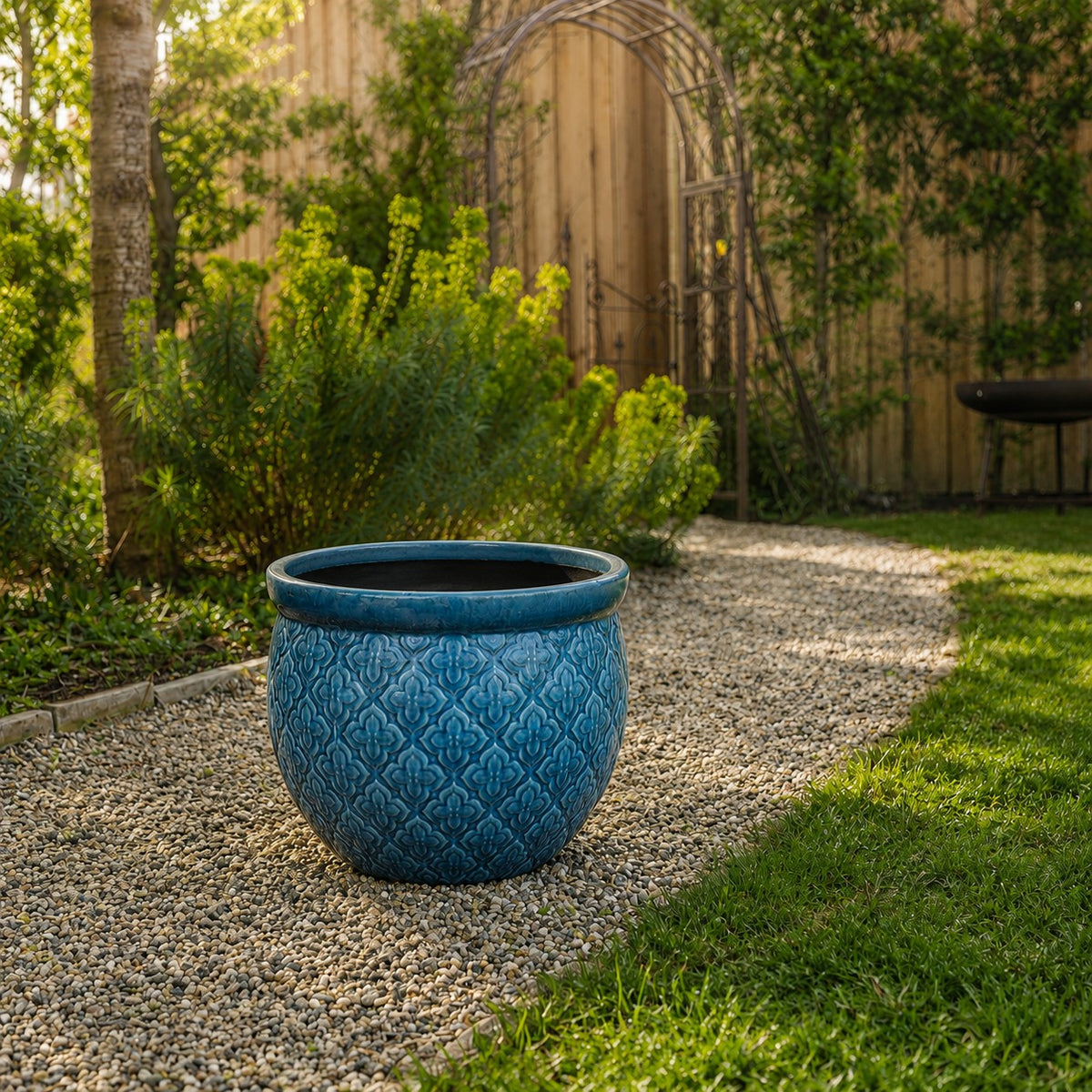 Ein großer blauer Keramikübertopf mit einem dekorativen Muster steht auf einem Kiesweg in einem sonnenbeschienenen Garten, umgeben von grünen Pflanzen und einem Holzzaun im Hintergrund.