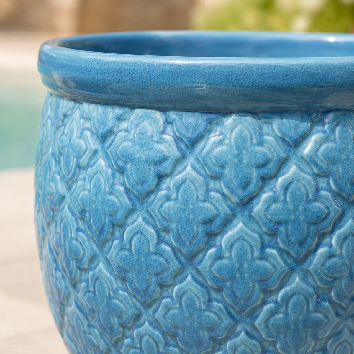 Nahaufnahme eines blauen Keramik-Pflanzgefäßes mit glänzender Oberfläche und strukturiertem, geometrischem Blumenmuster. Der Übertopf steht im Freien, mit einem unscharfen Hintergrund, der einen Pool und einen Terrassenbereich zeigt.