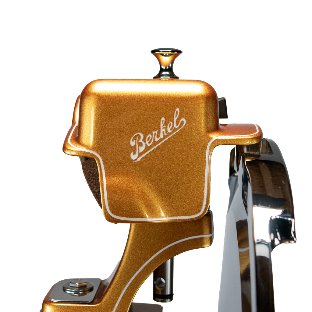 Nahaufnahme einer Berkel Volano B114 Schwungradmaschine Gold | mit Fassholz-Einlegebrett. Zu sehen sind die goldene Metallic-Oberfläche, das "Berkel"-Logo in weißer Schrift auf der Vorderseite, reflektierende Oberflächen und ein markanter Seitengriff.
