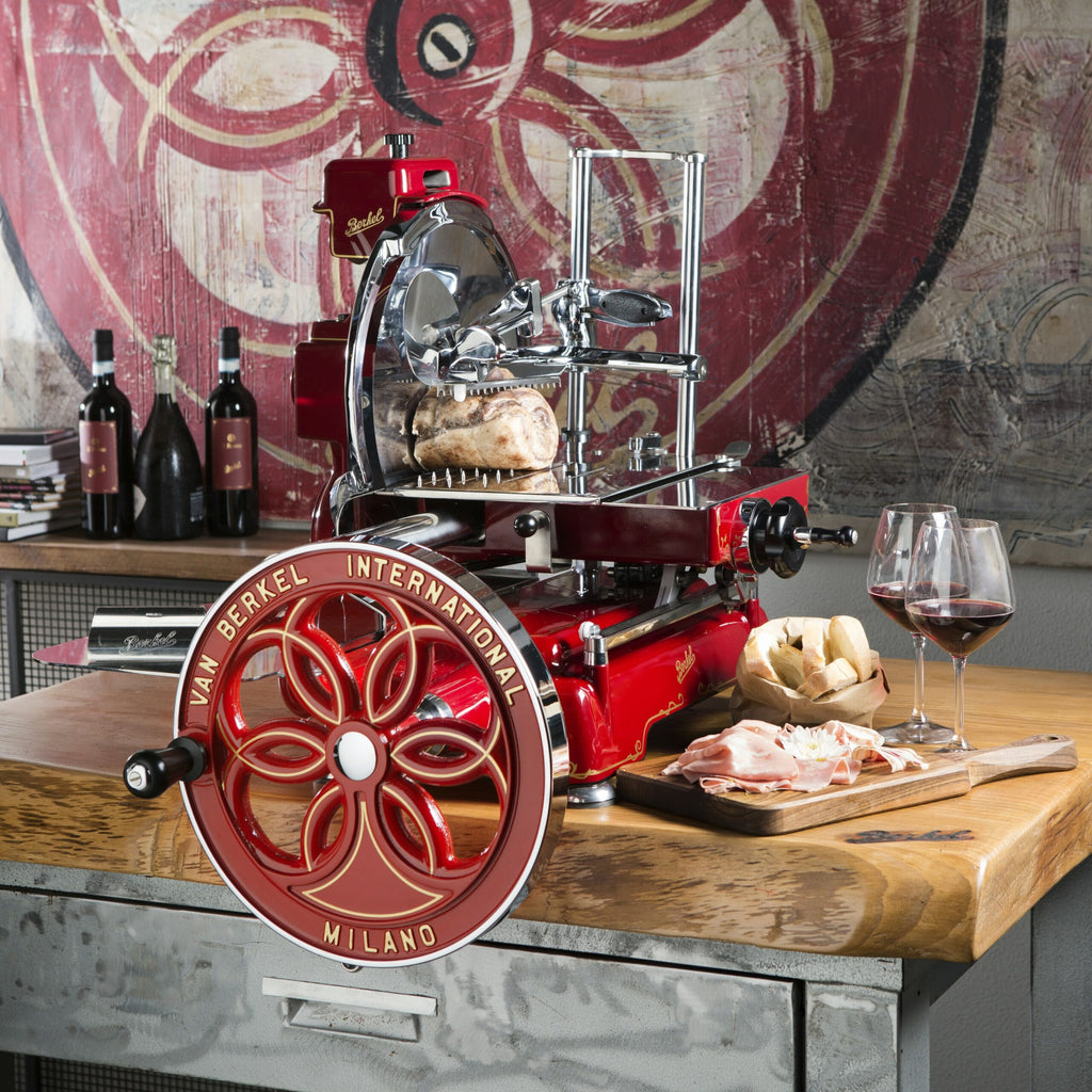 Eine lebhafte Berkel Volano B114 Rot manuelle Schwungradmaschine inkl. Standfuß steht auf einem Holztisch. Aufgeschnittenes Fleisch und Brot liegen auf einem Fassholzbrett, neben zwei gefüllten Weingläsern. Hinter der Aufschnittmaschine sind Weinflaschen und ein Buch auf einem Regal angeordnet, an der Wand hängt ein großes abstraktes Gemälde.