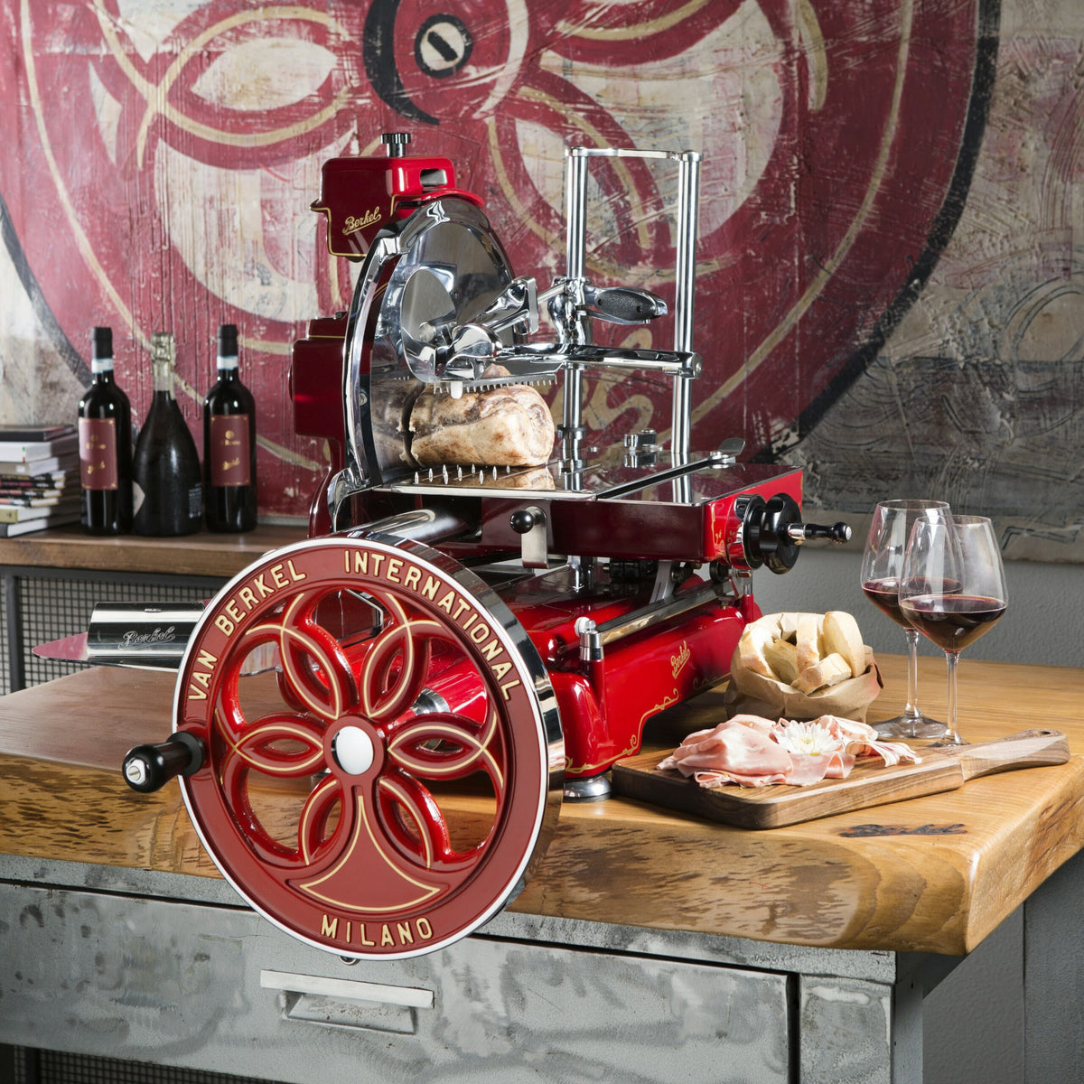 Eine lebhafte Berkel Volano B114 Rot manuelle Schwungradmaschine inkl. Standfuß steht auf einem Holztisch. Aufgeschnittenes Fleisch und Brot liegen auf einem Fassholzbrett, neben zwei gefüllten Weingläsern. Hinter der Aufschnittmaschine sind Weinflaschen und ein Buch auf einem Regal angeordnet, an der Wand hängt ein großes abstraktes Gemälde.