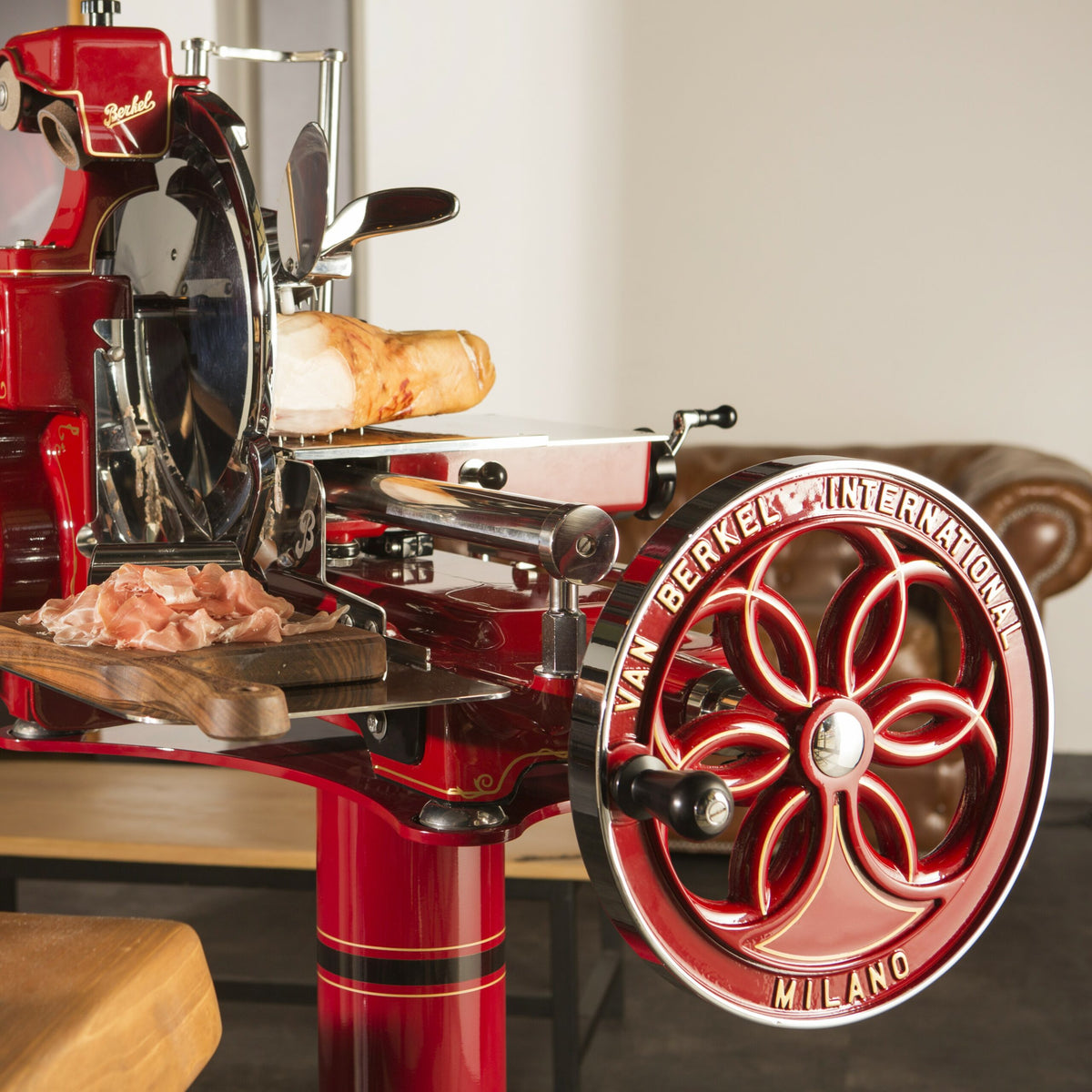 Eine Nahaufnahme einer alten Aufschnittmaschine für rotes Fleisch, der Berkel Volano B114 Rot manuelle Schwungradmaschine inklusive Standfuß und Fassholzbrett, mit dem Namen „Van Berkel International Milano“ auf dem Griffrad. Prosciuttoscheiben sind auf einem Fassholzbrett vor der Schwungradmaschine angeordnet und ein großes Stück Fleisch wird zum Schneiden geladen.