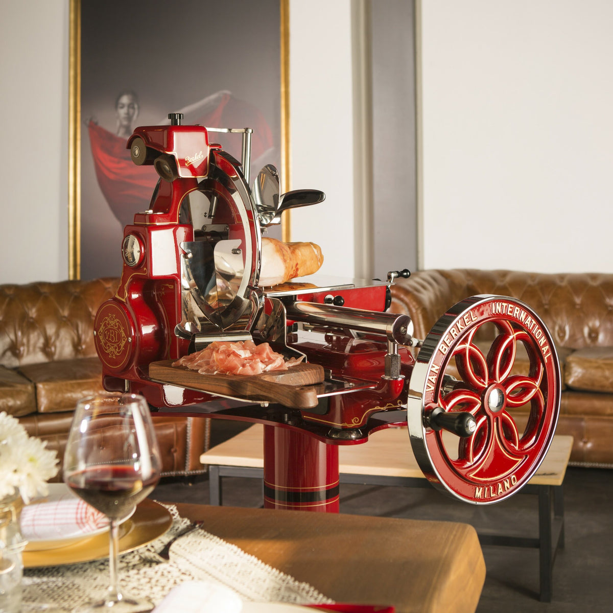 Eine alte Aufschnittmaschine für rotes Fleisch, genauer gesagt eine Berkel Volano B114 Rot manuelle Aufschnittmaschine mit passgenauen Einlegebrett, steht auf einem Tisch in einem gemütlichen Raum mit einem braunen gepolsterten Ledersofa und einem Porträt an der Wand. Die Aufschnittmaschine schneidet mühelos sowohl durch Brot als auch durch dünne Fleischscheiben. Ein Glas Rotwein und ein Teller mit Essen schmücken den Tisch im Vordergrund.