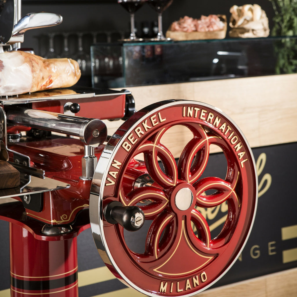 Eine Nahaufnahme einer manuellen Schwungradmaschine vom Typ Berkel Volano B114 Rot mit der Aufschrift „Van Berkel International Milano“ auf dem Vorderrad. Im Hintergrund sind Weingläser, ein Laib Brot und ein Bündel Kräuter auf einer Theke aus Fassholzbrett zu sehen.