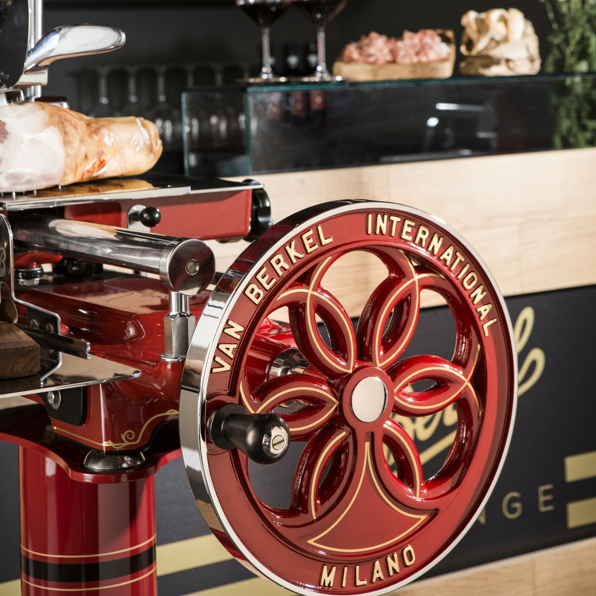 Eine Nahaufnahme einer manuellen Schwungradmaschine vom Typ Berkel Volano B114 Rot mit der Aufschrift „Van Berkel International Milano“ auf dem Vorderrad. Im Hintergrund sind Weingläser, ein Laib Brot und ein Bündel Kräuter auf einer Theke aus Fassholzbrett zu sehen.