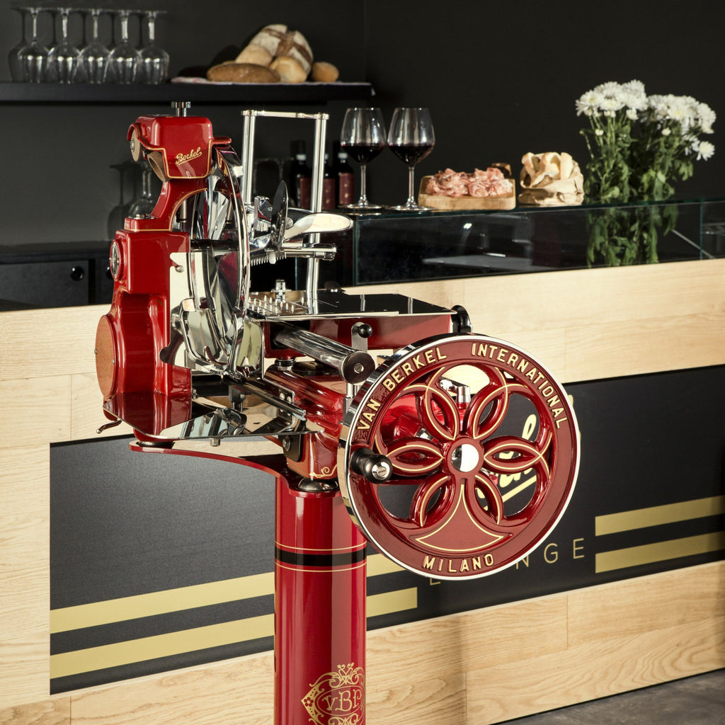 Eine Berkel Volano B114 Rot manuelle Schwungradmaschine inkl. Standfuß + Fassholzbrett wird in einem stilvollen, modernen Feinkostladen ausgestellt. Die manuelle Fleischschneidemaschine mit der Aufschrift „Van Berkel International Milano“ auf dem Rad steht vor einer Theke mit Weingläsern, einer Blumenvase und verschiedenen Brotsorten und Fleischsorten im Hintergrund.