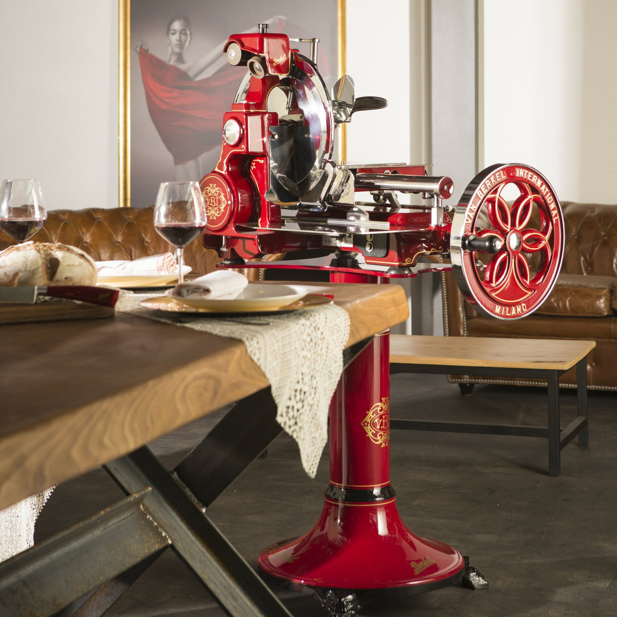 Eine leuchtend rote, im Vintage-Stil gehaltene Berkel Volano B114 Rot manuelle Schwungradmaschine inkl. Standfuß + Fassholzbrett-Fleischschneidemaschine steht prominent auf einem Holztisch, der mit weißen Spitzentischsets, Weingläsern und Tellern geschmückt ist. Im Hintergrund sind ein braunes Ledersofa und ein gerahmtes Porträt einer Frau in einem roten Kleid zu sehen.