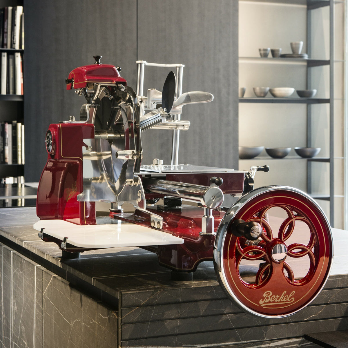 Eine alte Berkel Volano B3 Rot Schwungradmaschine mit Standfuß und Fassholzeinlegebrett steht in einer modernen Küche auf einer Marmorarbeitsplatte. Die Aufschnittmaschine, verziert mit aufwendigen Verzierungen und dem Berkel-Logo auf dem Rad, steht stolz da. Im Hintergrund sind Regale mit Schüsseln und ein Bücherregal zu sehen.