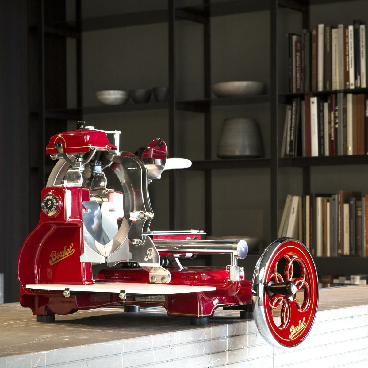 Eine alte Berkel Volano B3 Rot Schwungradmaschine mit Standfuß und Fassholzeinlegebrett mit polierten Metalldetails steht auf einer Küchentheke. Im Hintergrund sind Regale voller Bücher und dekorative Töpferwaren zu sehen, die auf eine stilvolle und moderne Kücheneinrichtung hindeuten.