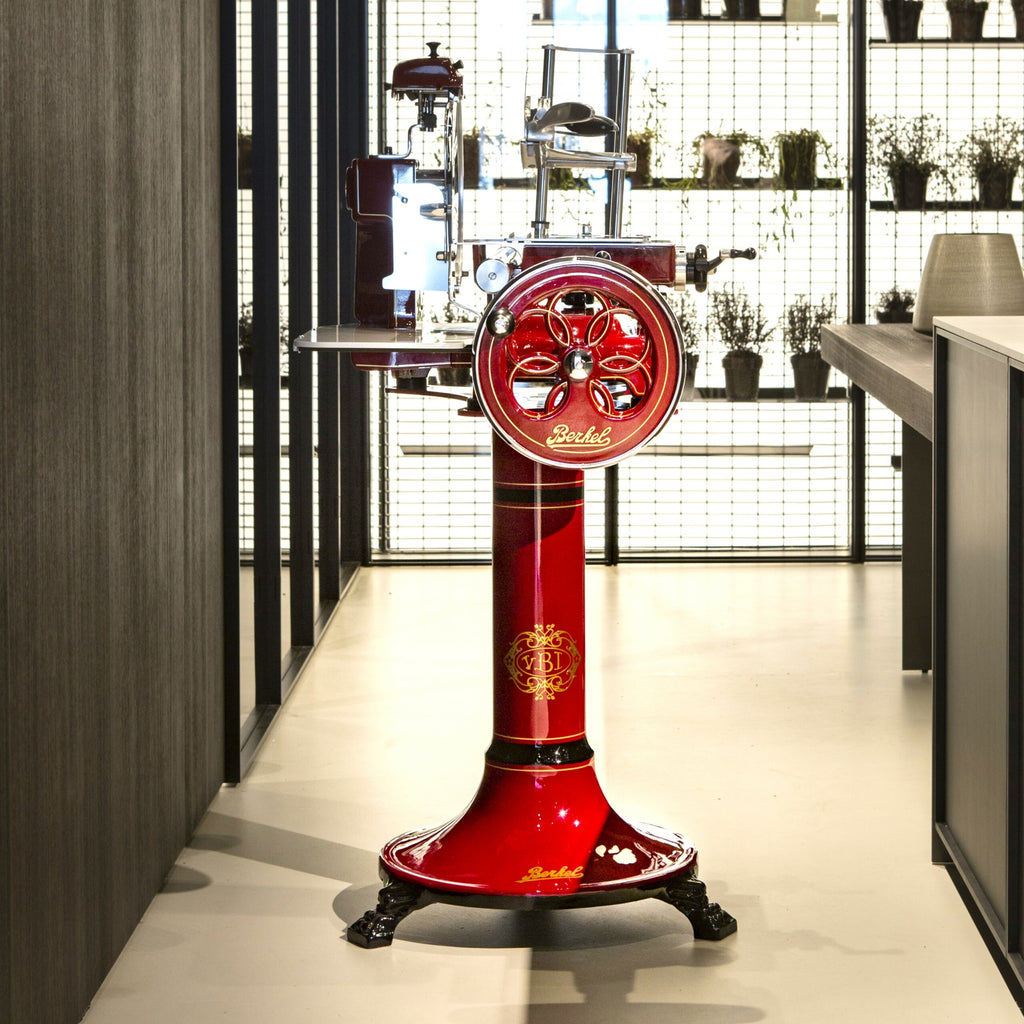 Eine Berkel Volano B3 Rot Schwungradmaschine mit Standfuß und Fassholzeinlegebrett steht auf einem polierten Boden in einem modernen Raum mit dunklen Wänden. Der Allesschneider hat ein kunstvolles Design, komplett mit einem Handkurbelrad und metallischen Akzenten. Im Hintergrund sind Regale mit Topfpflanzen und Küchenutensilien geschmückt, die von hellem Licht erhellt werden.