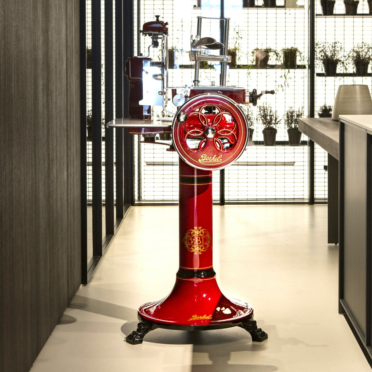 Eine Berkel Volano B3 Rot Schwungradmaschine mit Standfuß und Fassholzeinlegebrett steht auf einem polierten Boden in einem modernen Raum mit dunklen Wänden. Der Allesschneider hat ein kunstvolles Design, komplett mit einem Handkurbelrad und metallischen Akzenten. Im Hintergrund sind Regale mit Topfpflanzen und Küchenutensilien geschmückt, die von hellem Licht erhellt werden.