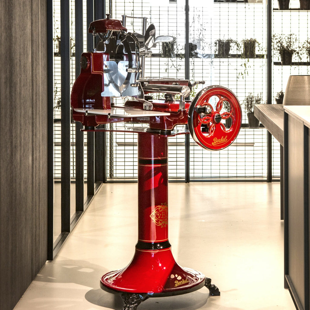 Eine Vintage-Berkel Volano B3 Rot, Schwungradmaschine mit Standfuß und Fassholzeinlegebrett steht auf einem glänzenden Sockel in einer modernen Küchenumgebung. Der Allesschneider ist mit kunstvollen Details und einem Handkurbelrad versehen. Im Hintergrund steht ein Gitterregal mit verschiedenen Topfpflanzen, das dem schlichten, industriellen Dekor einen Hauch von Grün verleiht.
