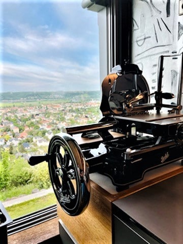 Eine Vintage-Schneidemaschine steht neben einem großen Fenster, durch das man eine weitläufige Landschaft mit einer entfernten Stadt und grünen Feldern sehen kann. Terrakotta-Töpfe auf der Fensterbank sorgen für rustikalen Charme, während die Metallteile der Maschine im Tageslicht glitzern und eine Mischung aus industrieller und ruhiger Außenlandschaft schaffen.
