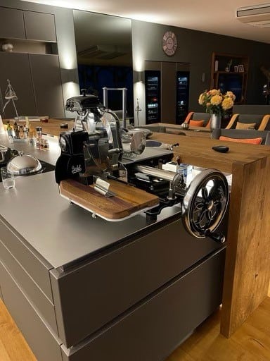 Eine elegante, moderne Küche mit einer großen Kochinsel und einem Fleischschneider im Vintage-Stil. Die Arbeitsflächen sind sauber und die verschiedenen Küchenutensilien sind ordentlich angeordnet. Im Hintergrund sorgen neben Barhockern und einer Vase mit Blumen Amphoren- und Terrakotta-Töpfe neben der Wanduhr für rustikalen Charme.