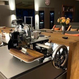 Eine elegante, moderne Küche verfügt über eine Berkel Volano B3 Schwarz Aufschnittmaschine mit Schwungrad sowie ein Aufschnittbrett aus Fassholz, das auf der Arbeitsplatte montiert ist. Im Hintergrund stehen Barhocker mit orangefarbenen Kissen, ein Blumenstrauß und verschiedene Küchengeräte vor stilvollen, dunklen Schränken.