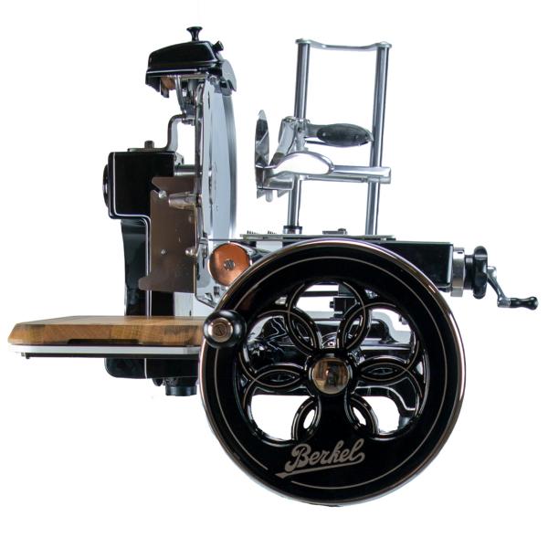 Ein Berkel Volano B3 Schwarz Fleischschneider im Vintage-Stil mit Chromakzenten von der Seite betrachtet. Er verfügt über einen Schwungradmechanismus mit Handkurbel und aufwendigem Raddesign, eine Holzplattform aus Fassholz zum Platzieren des Fleisches und ein glattes, poliertes Gehäuse.
