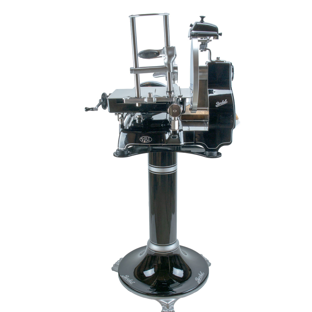 Eine alte Berkel Volano B3 Schwarz Schwungrad-Aufschnittmaschine in schwarz-silbernem Design verfügt über eine Handkurbel, die auf einem zylindrischen Fassholz-Ständer mit integriertem Schneidebrett montiert ist. Diese Aufschnittmaschine verfügt über ein rundes Messer und verschiedene mechanische Komponenten, wobei der Markenname „Berkel“ auf mehreren Teilen deutlich sichtbar angebracht ist.