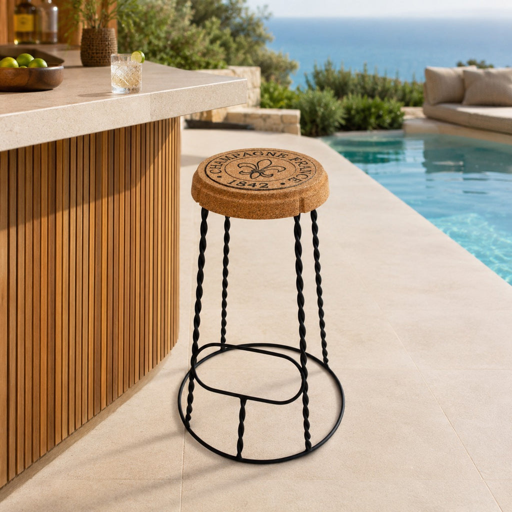 Der hochwertige Barhocker aus Kork und Metall "Agraffe" mit gedrehten schwarzen Beinen und Sitzfläche mit Stempel und Fleur-de-Lis-Design steht neben einer Poolbar, umgeben von Pool, Grünpflanzen und Meerblick.