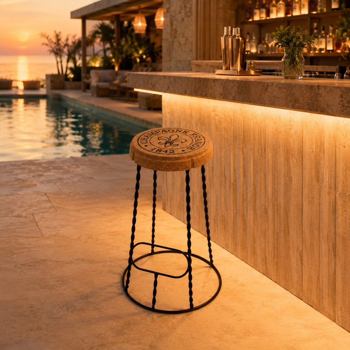 Der hochwertige Barhocker aus Kork und Metall "Agraffe", inspiriert von einem Tequila-Flaschendeckel, steht bei Sonnenuntergang an einer Steinbar am Pool, umgeben von Drinks und üppigen Pflanzen.