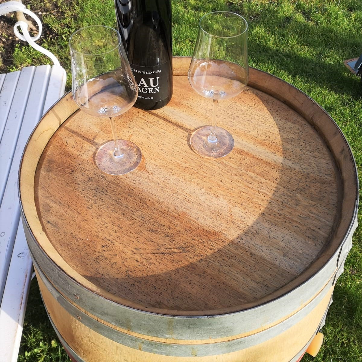 Ein Original Weinfass aus Eiche, Barriquefass 225 Liter, gefüllt mit einer Flasche Wein und zwei leeren Weingläsern, steht an einem sonnigen Platz im Freien. Das Fass dient als provisorischer Tisch auf grünem Gras neben einer weißen Bank. Schatten deuten auf einen hellen und klaren Tag hin.