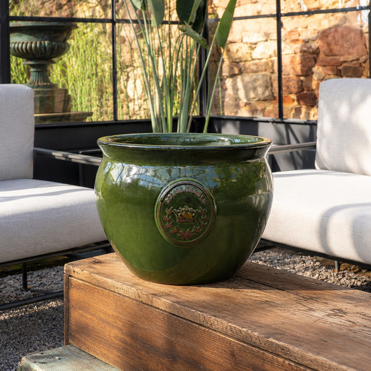 Der bauchige glasierte Pflanztopf "Royal Garden Green" (Ø 16-38cm) steht auf einem rustikalen Holztisch in einer sonnigen, modernen Outdoor-Sitzecke mit gepolsterten Stühlen und üppigem Blattwerk im Hintergrund.
