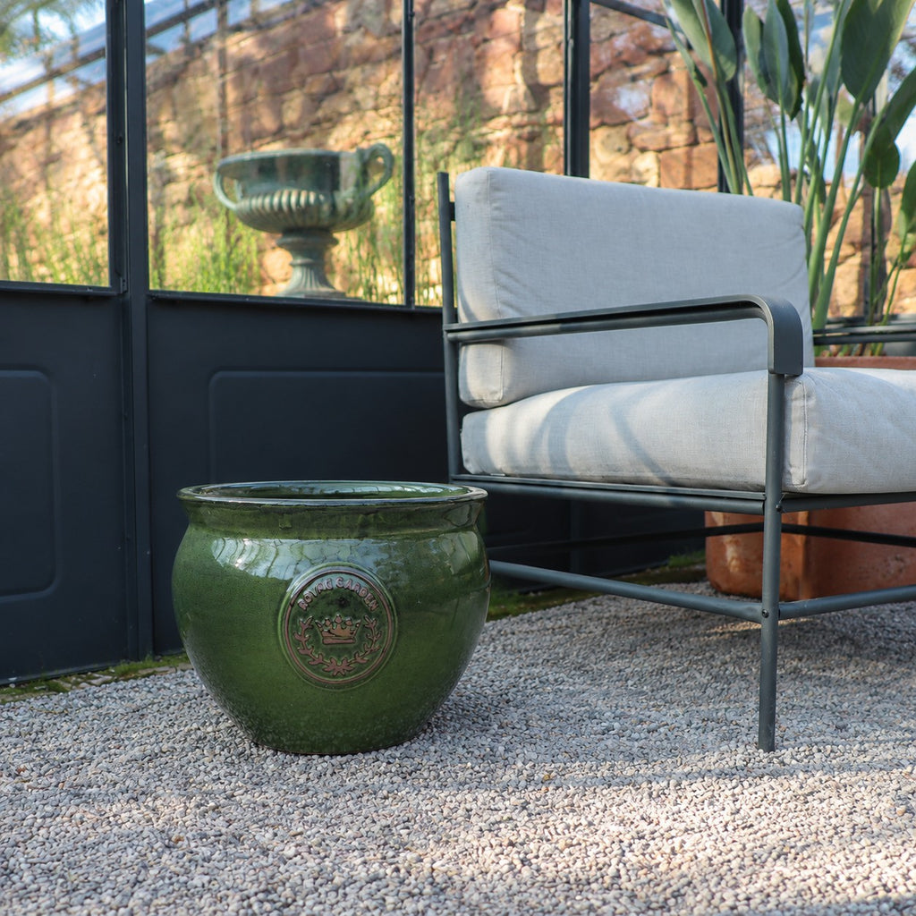 Der bauchige glasierte Pflanztopf "Royal Garden Green" (Durchmesser 16-38cm) sitzt auf Kies neben einem modernen grauen gepolsterten Sessel in einem Raum mit Glaswänden, mit Pflanzen und einer Steinurne im Hintergrund.