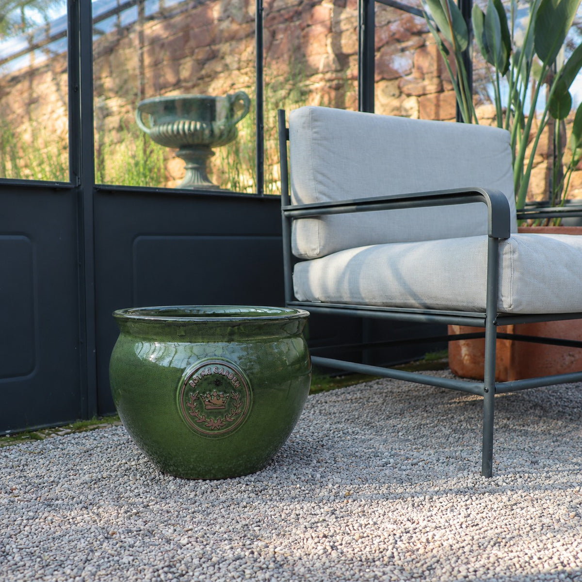 Der bauchige glasierte Pflanztopf "Royal Garden Green" (Durchmesser 16-38cm) sitzt auf Kies neben einem modernen grauen gepolsterten Sessel in einem Raum mit Glaswänden, mit Pflanzen und einer Steinurne im Hintergrund.