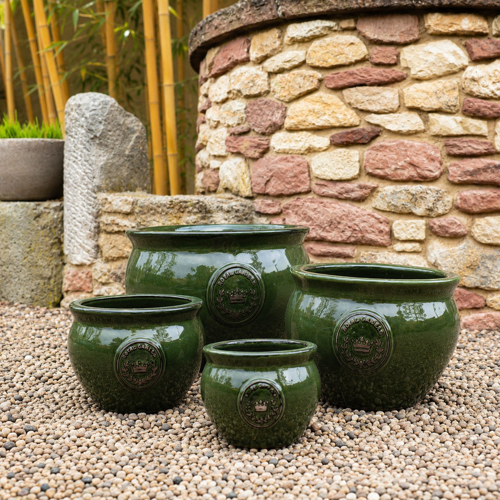 Fünf "Royal Garden Green" bauchiger glasierter Pflanztopf in dunkelgrün, 16-38cm Durchmesser, mit dekorativen Emblemen sind auf Kies im Freien vor einer Steinmauer und Bambus angeordnet.