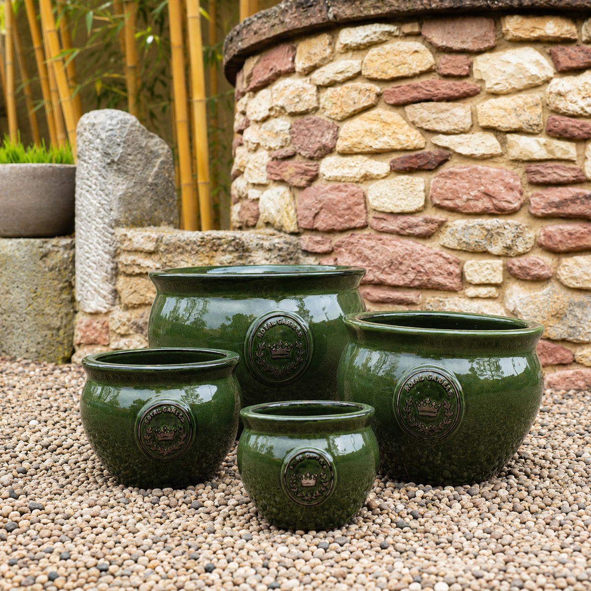Fünf "Royal Garden Green" bauchiger glasierter Pflanztopf in dunkelgrün, 16-38cm Durchmesser, mit dekorativen Emblemen sind auf Kies im Freien vor einer Steinmauer und Bambus angeordnet.