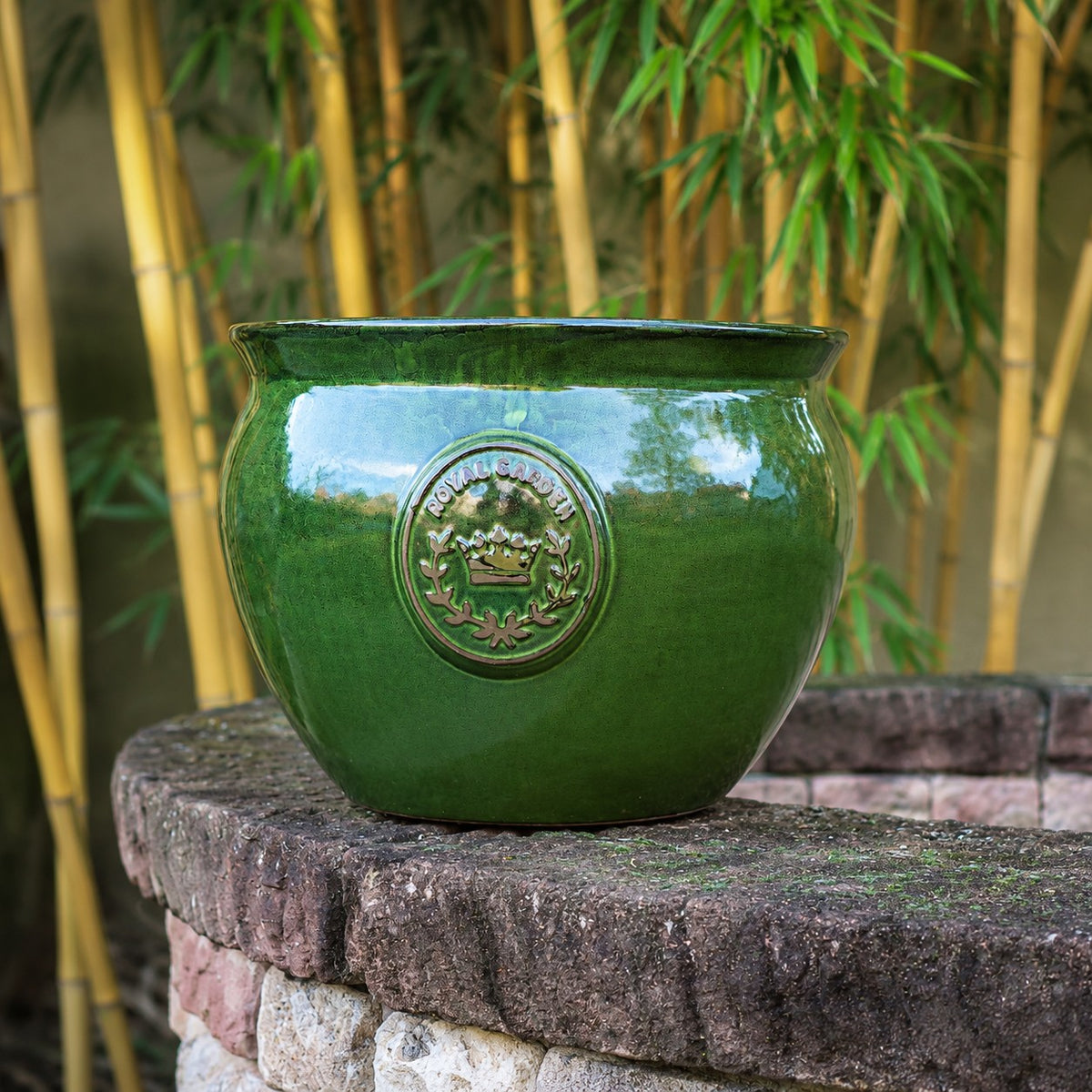 Der bauchige glasierte Pflanztopf "Royal Garden Green" (Ø 16-38cm) mit geprägtem Emblem sitzt auf einer runden Steinmauer, eingerahmt von hohen Bambuspflanzen im Hintergrund.