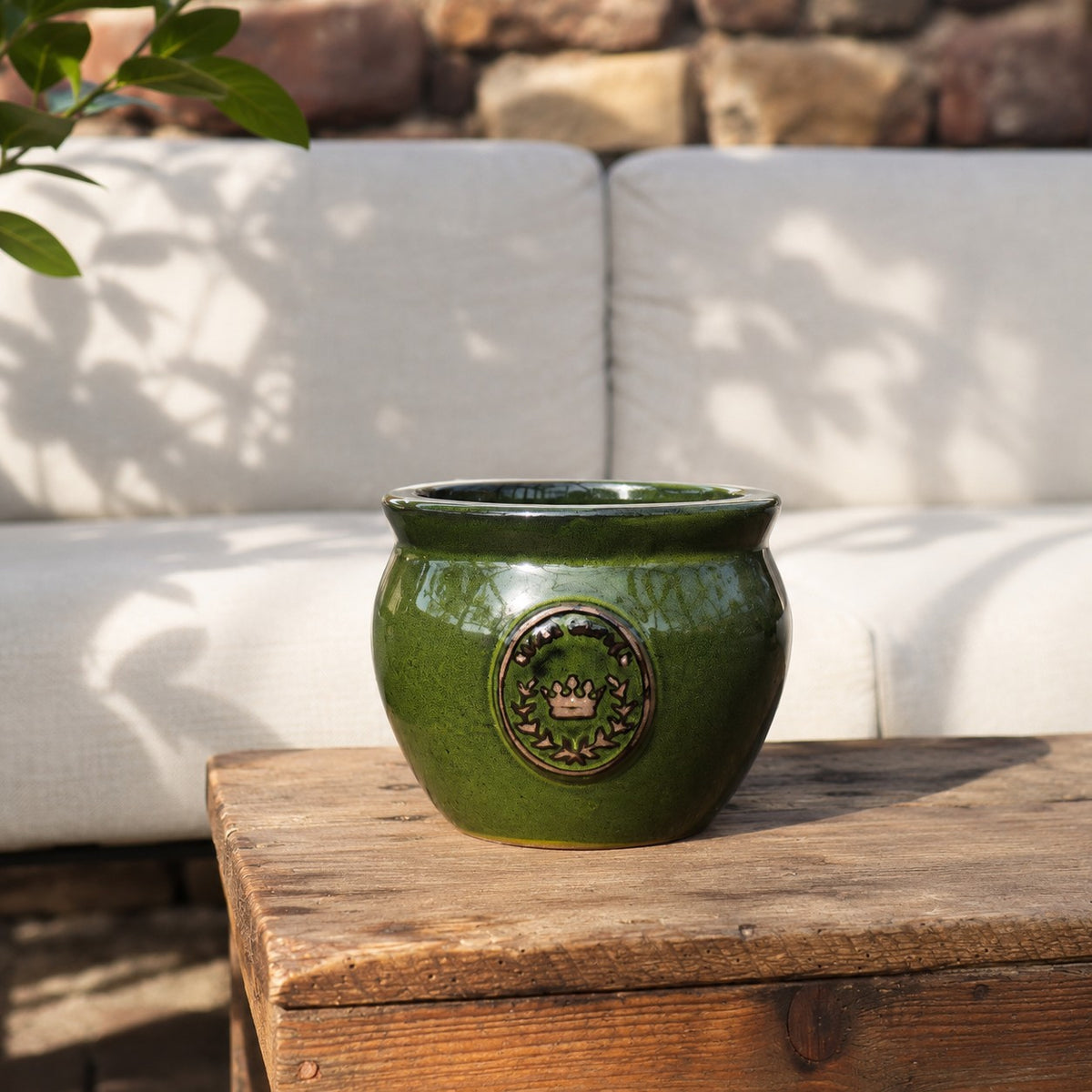 Der bauchige glasierte Pflanztopf "Royal Garden Green" (Ø 16-38 cm) glänzt auf einem rustikalen Holztisch im Freien, vor einem weiß gepolsterten Sofa und einer Steinwand, während Sonnenlicht blättrige Schatten wirft.