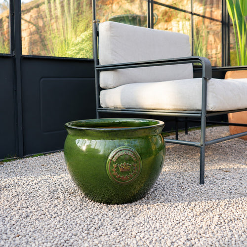 Der bauchige glasierte Pflanztopf "Royal Garden Green" (Durchmesser 16-38cm) mit dekorativem Emblem steht auf Kies vor einem modernen Outdoor-Stuhl mit leichten Kissen in einem sonnendurchfluteten Glasraum.