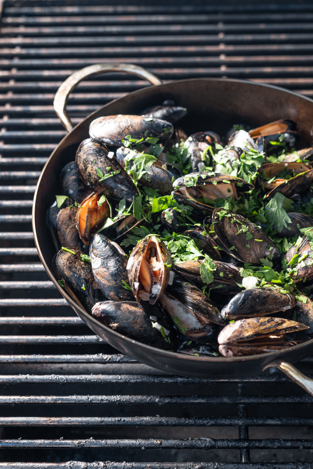 A De Buyer Bauerntopf Mineral B mit zwei Griffen, gefüllt mit gekochten Muscheln und frischen Kräutern garniert, steht auf einem Grill mit Metallrost.