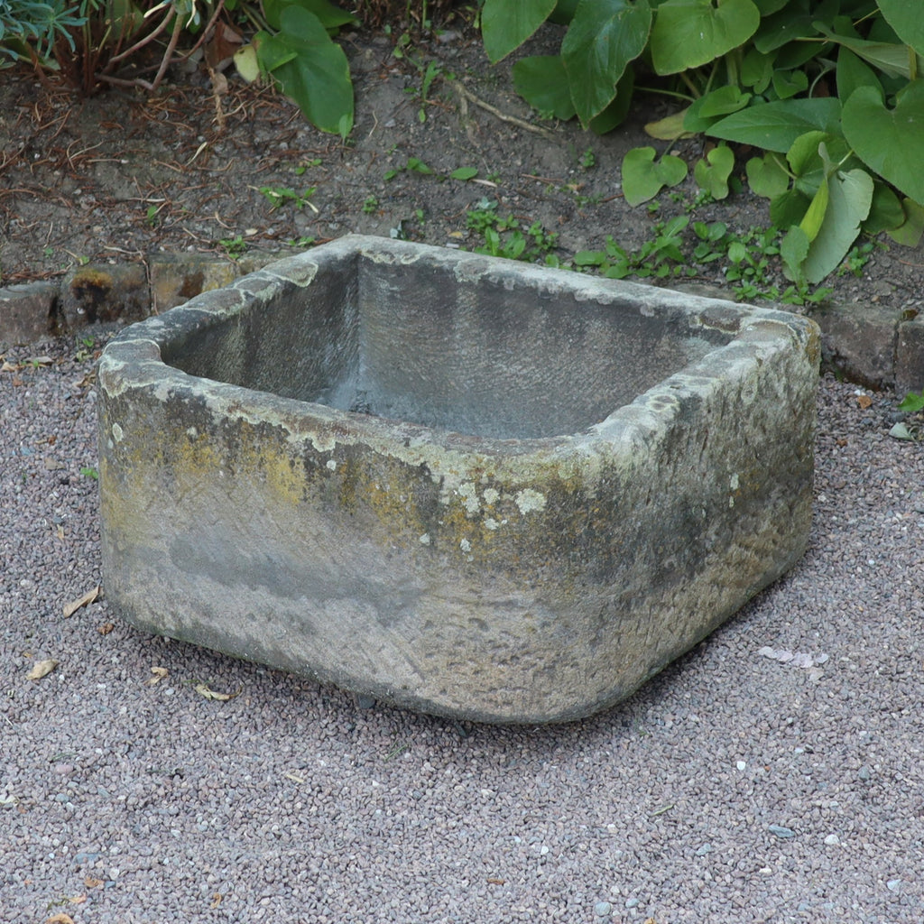 Ein altertümliches Sandsteinbecken 65 x 61 x 31 cm steht auf Kies, umrahmt von Ziegelsteinen und niedrigen Grünpflanzen, und bildet eine reizvolle Gartenszene.