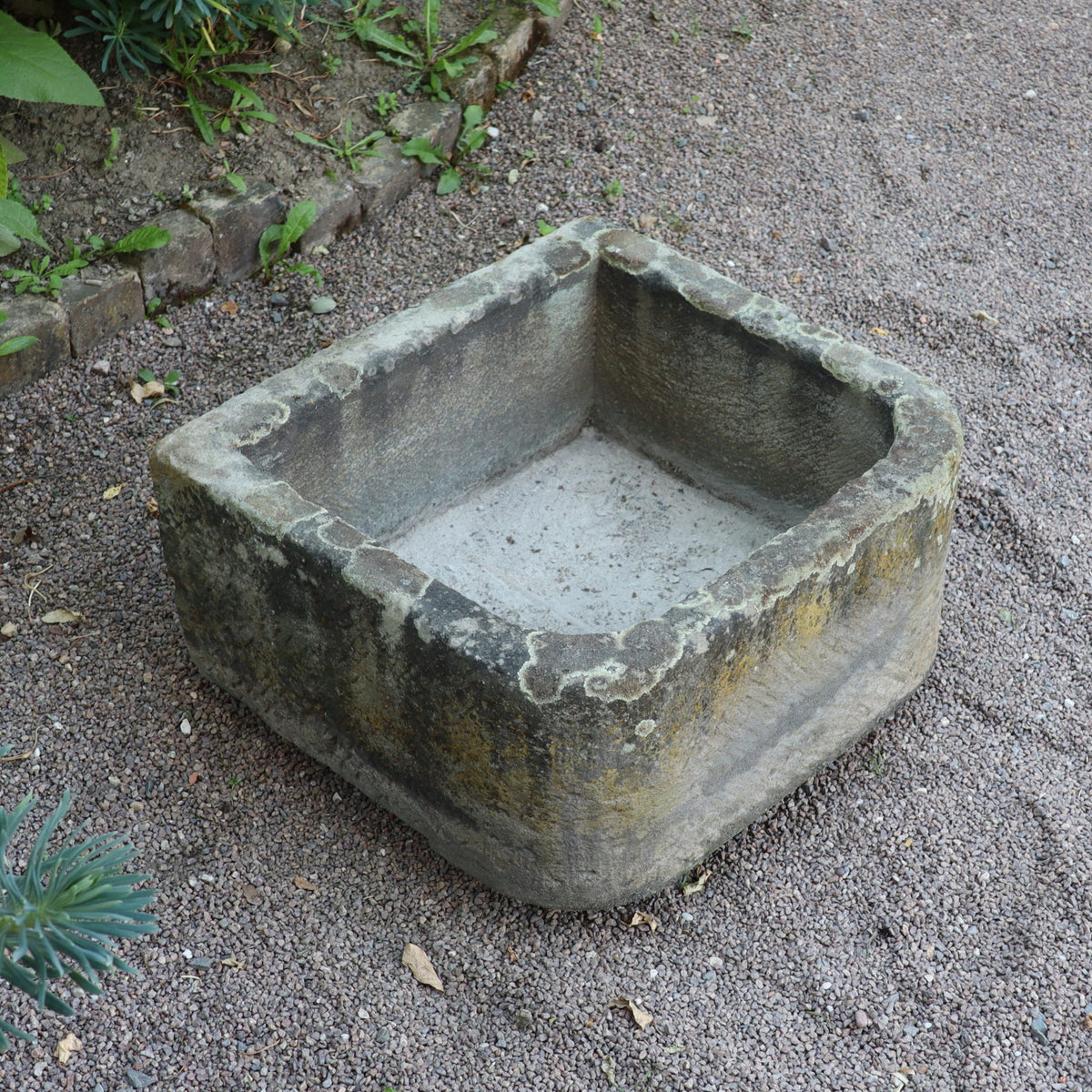 Ein altertümliches Sandsteinbecken 65 x 61 x 31 cm mit verwitterten Kanten ruht auf Kies neben einem üppigen Gartenbeet.