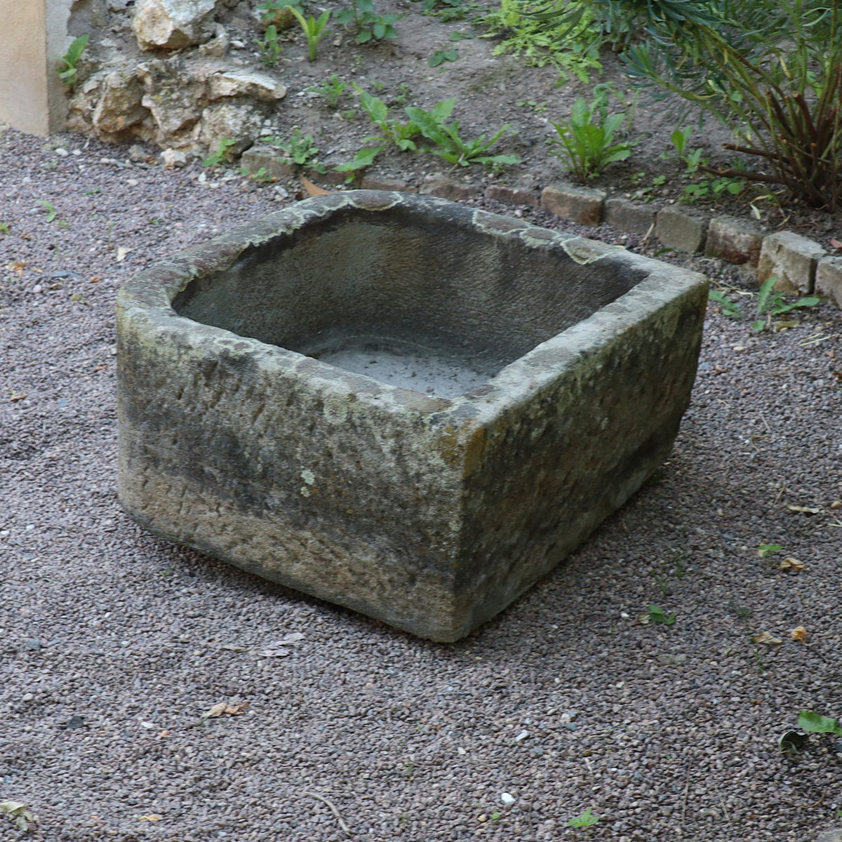 Ein altes Sandsteinbecken (65 x 61 x 31 cm) liegt auf Kies in einem Garten, umgeben von Pflanzen und eingefasst von Ziegeln und Erde.