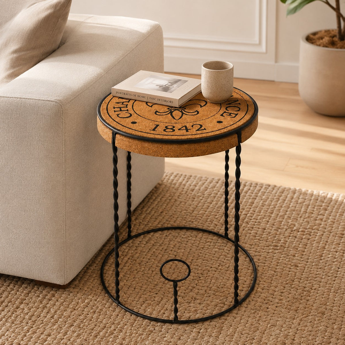 Der Hochwertige Beistelltisch aus Kork "Agraffe" mit einer Korkplatte und schwarzen Metallbeinen steht neben einem beigen Sofa mit einem Buch und einer Keramiktasse auf einem gewebten Teppich über Holzboden neben einer Topfpflanze.