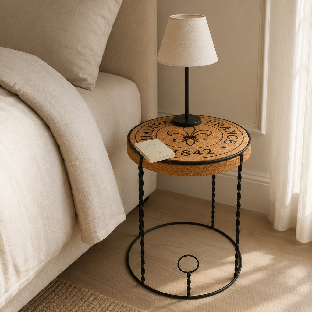 Eine gemütliche Nachttischszene mit einem Bett in beiger Bettwäsche, einem hochwertigen Beistelltisch aus Kork "Agraffe" mit französischem Text, einer kleinen beigen Lampe und einem aufgeschlagenen Buch, während das Sonnenlicht durch die transparenten Vorhänge fällt.