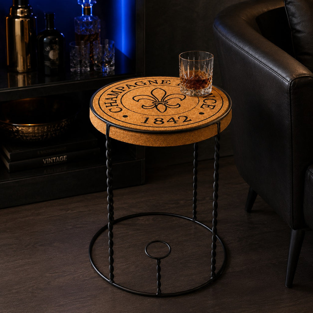 Ein Hochwertiger Beistelltisch aus Kork "Agraffe" mit runder Platte, fleur-de-lis und "CHAMPAGNE FRANCE 1842" steht neben einem schwarzen Ledersessel. Darauf ein Glas Whiskey, im Hintergrund ein beleuchtetes Barregal.