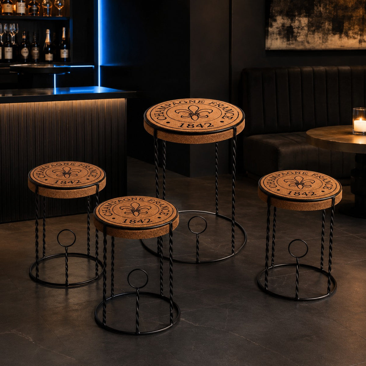 Vier Barhocker mit Sitzflächen aus Kork, inspiriert vom Beistelltisch "Agraffe", sind mit dem Schriftzug "CHAMPAGNE PERRIER 1842" und einem Fleur-de-Lis-Motiv auf robusten Metallfüßen versehen, perfekt für eine moderne Lounge.