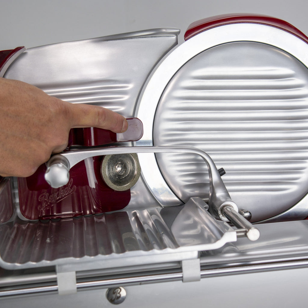 Nahaufnahme einer Hand, die einen Hebel an einer Fleischschneidemaschine im Vintage-Stil mit metallisch glänzender Oberfläche und roten Akzenten verstellt, mit dem Schleifaufsatz für Berkel Aufschnittmaschine Home Line 200 oder 250 + Poliertuchaufsatz.