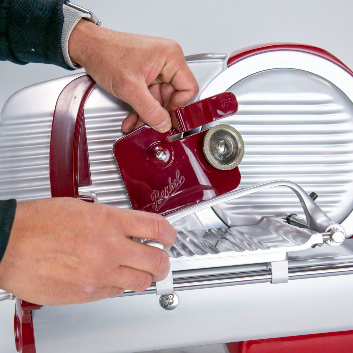 Nahaufnahme einer Person, die eine Berkel Home Line 200-Fleischschneidemaschine im Vintage-Stil mit rotem und silbernem Design einstellt. Der Markenname „Berkel“ ist auf dem roten Teil zu sehen, das eingestellt wird. Die Person, die ein schwarzes Langarmhemd und eine Uhr am linken Handgelenk trägt, stellt die Feineinstellung der Schneidemaschine mithilfe eines Schleifaufsatzes aus dem Berkel Home Line 200-Zubehörset ein, das Messerabnehmer, Abdeckung und Poliertuch umfasst.