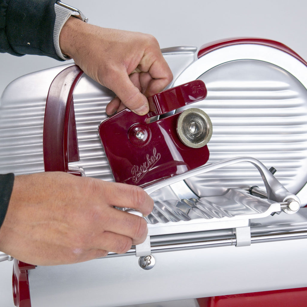Eine Person stellt eine rot-silberne Aufschnittmaschine mit dem Schleifaufsatz für Berkel Aufschnittmaschine Home Line 200 oder 250 + Poliertuch auf einer grauen Fläche ein und hält den seitlichen Knopf und den Hebel fest.