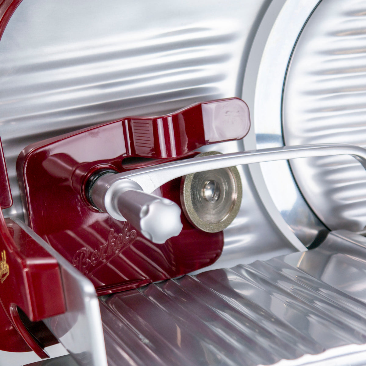 Nahaufnahme eines Vintage-Zubehörsets für die Berkel Home Line 200, bestehend aus Messerabnehmer, Schleifaufsatz, Abdeckung und Poliertuch, mit rotem Griff und markantem Rundmesser. Die Metallteile und die glänzende Oberfläche reflektieren das Licht hervorragend und betonen das komplizierte Design und die mechanischen Teile. Der Markenname ist auf dem roten Abschnitt teilweise sichtbar.