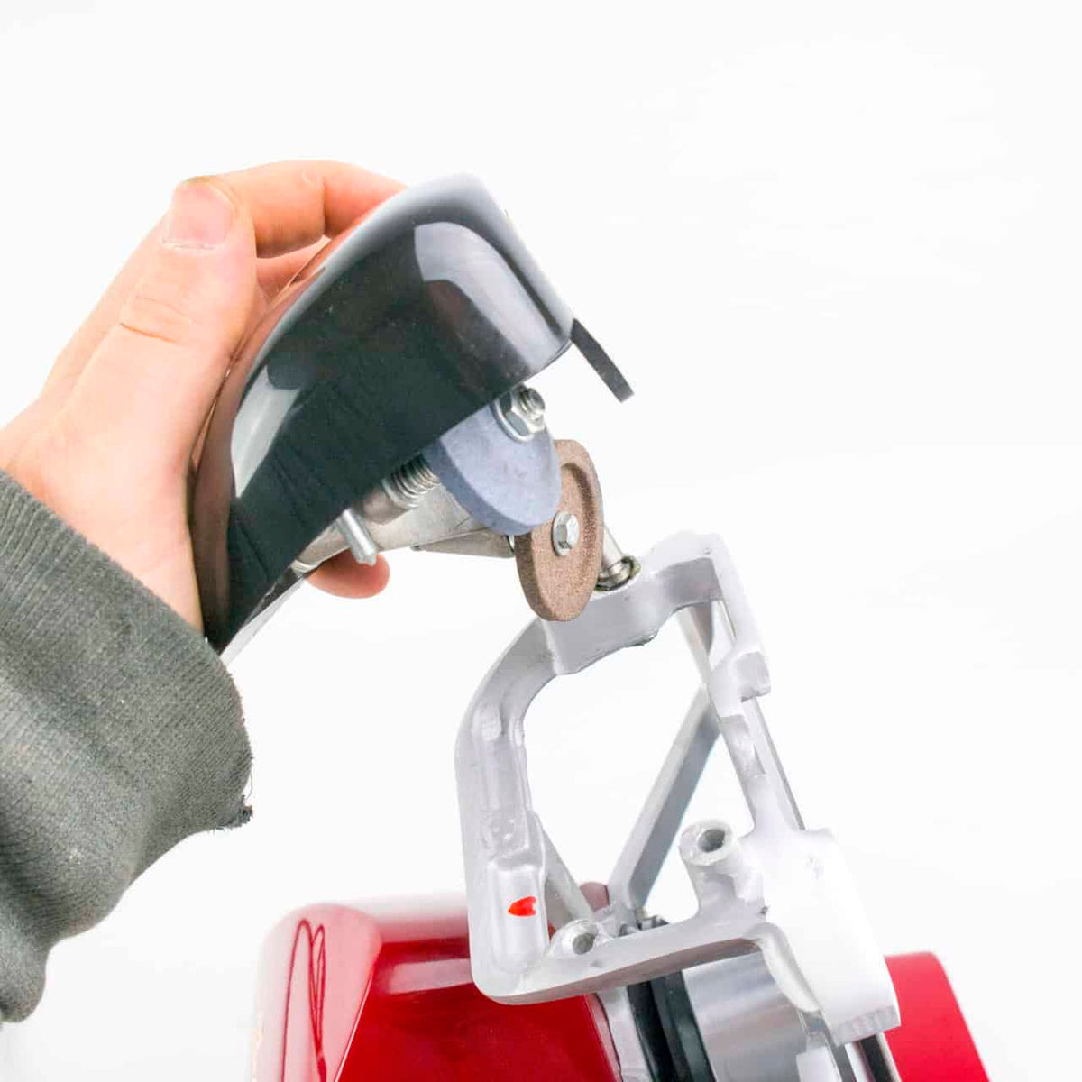 Die Hand einer Person hält ein schwarzes Gehäuse, das teilweise eine runde Schleifscheibe bedeckt, die an einem metallischen Werkzeug befestigt ist. Das Werkzeug scheint zum Schärfen oder Schleifen zu dienen und hat eine rote Basis, die dem Berkel Red Line 250 Rot Allesschneider ähnelt. Der Hintergrund ist schlicht weiß.