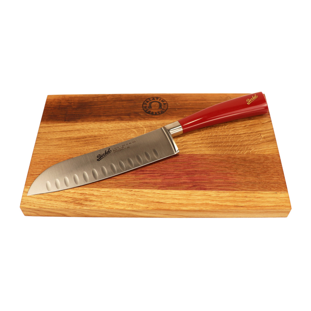 Ein Berkel Messer - Elegance Santoku-Messer mit rotem Griff ruht auf einem 100 % handgefertigten Schneidebrett Unikat mit den Maßen 30 x 18 cm. Die 18 cm lange Klinge, eine Anspielung auf traditionelles japanisches Design, weist eine Reihe von Einkerbungen auf und der Markenname Berkel ist in der Nähe des Griffs eingraviert. Das Schneidebrett weist in der oberen linken Ecke ein dekoratives Emblem auf.