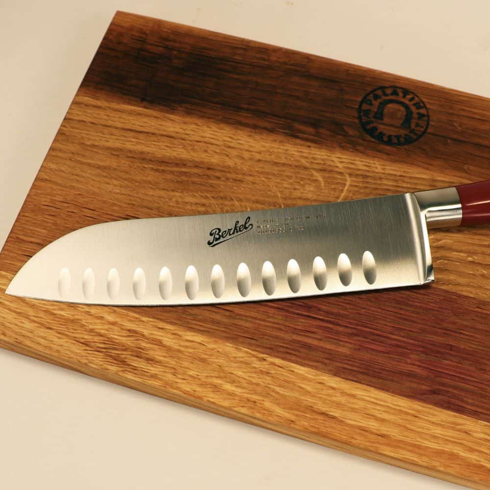 Ein 18 cm langes Santokumesser im japanischen Stil Elegance mit Granton-Schneide und rotem Griff liegt auf einem einzigartigen, 100 % handgefertigten Holzschneidebrett mit den Maßen 30 x 18 cm. In die Klinge des Messers ist der Markenname „Berkel Messer“ eingraviert. Das Schneidebrett hat in der Ecke ein kreisförmiges Emblem, das sein elegantes Aussehen unterstreicht.